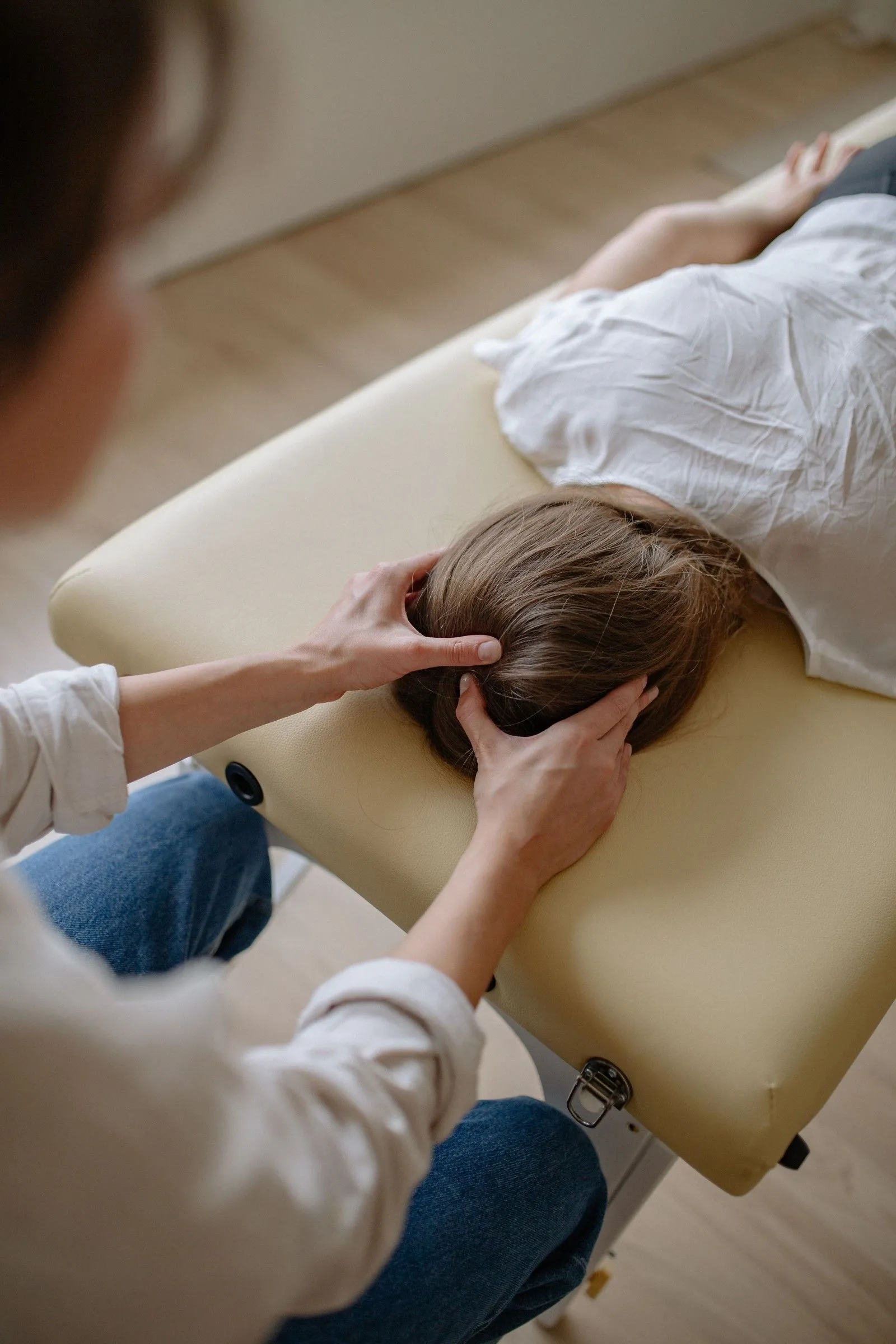 Seen from above in a daylit treatment room, a chiropractor's hands support the back of a patient's head as she lies face down