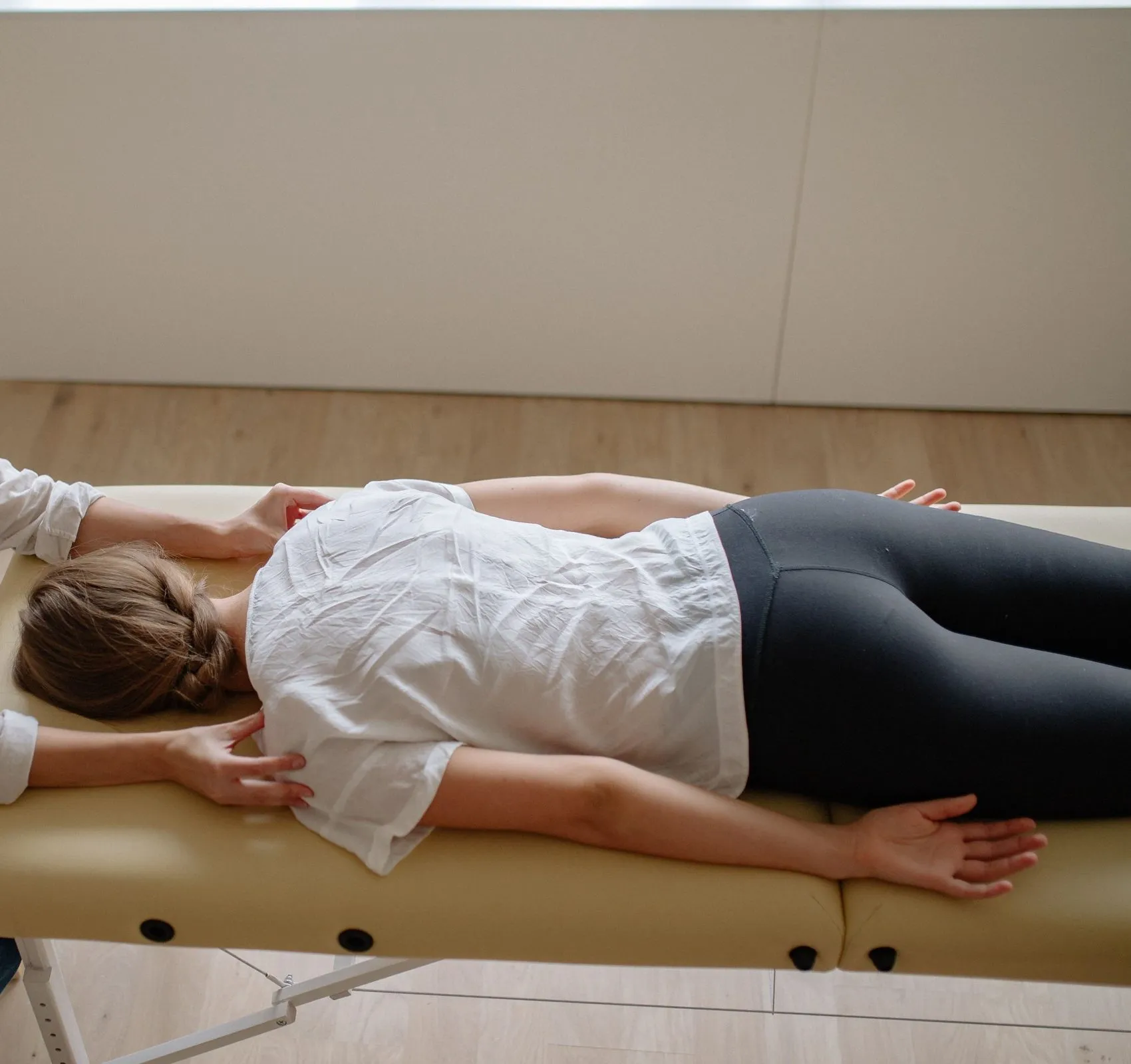 A patient lies face down on a cream treatment table in a bright clinic room while a chiropractor works on her shoulders